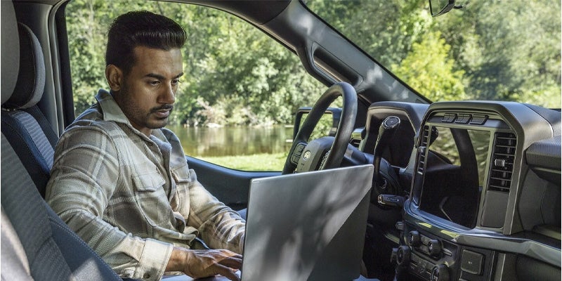 A man works on a laptop in the driver's seat of a car, parked facing a lake and trees.