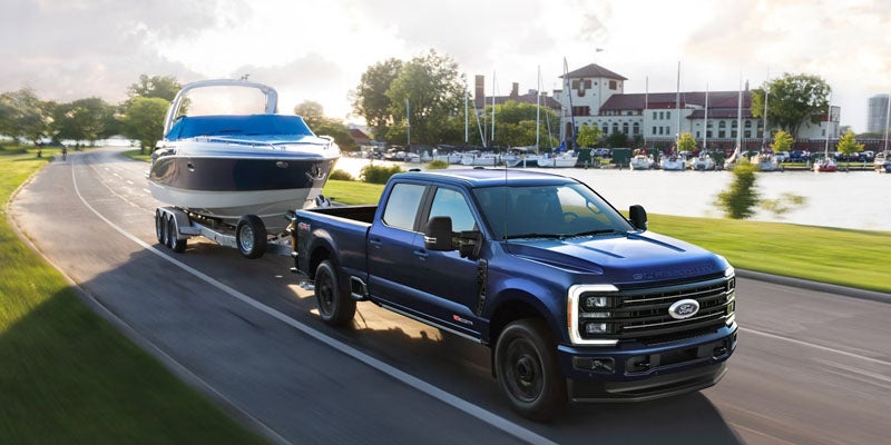 Blue Ford Super Duty dually pickup truck towing a livestock trailer across a grassy rural field, with a cowboy walking beside the trailer.